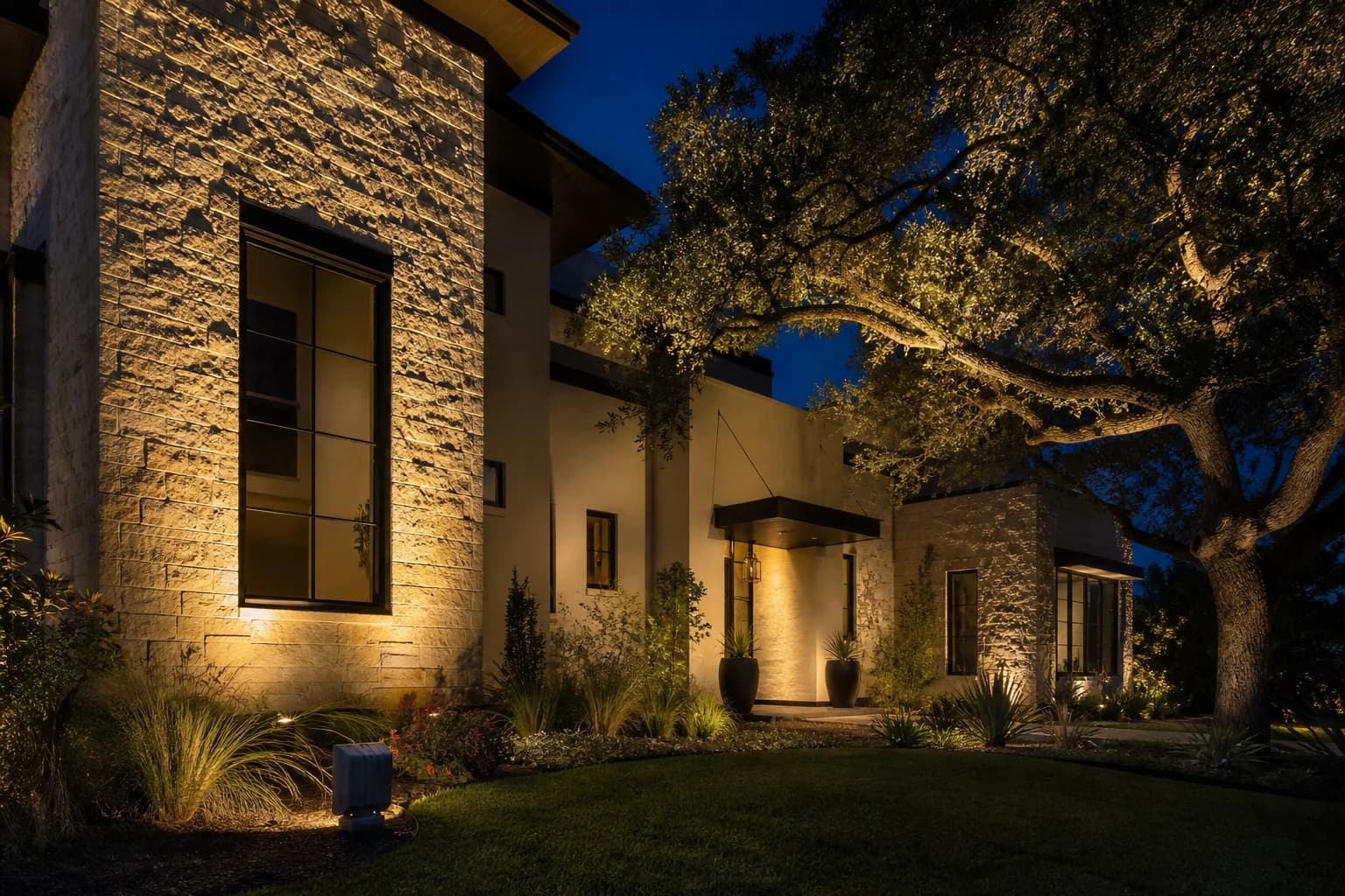 Large outdoor spotlight washing warm-white light across a fictional luxury home's limestone facade and live oak
